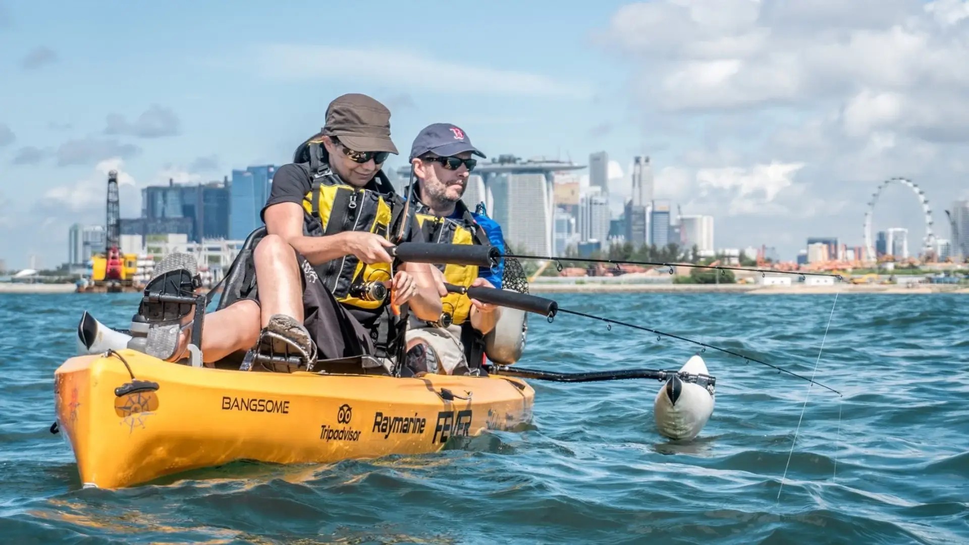 Two people kayak fishing off Sentosa with views of the Singapore skyline and Marina Bay Sands in the distance.