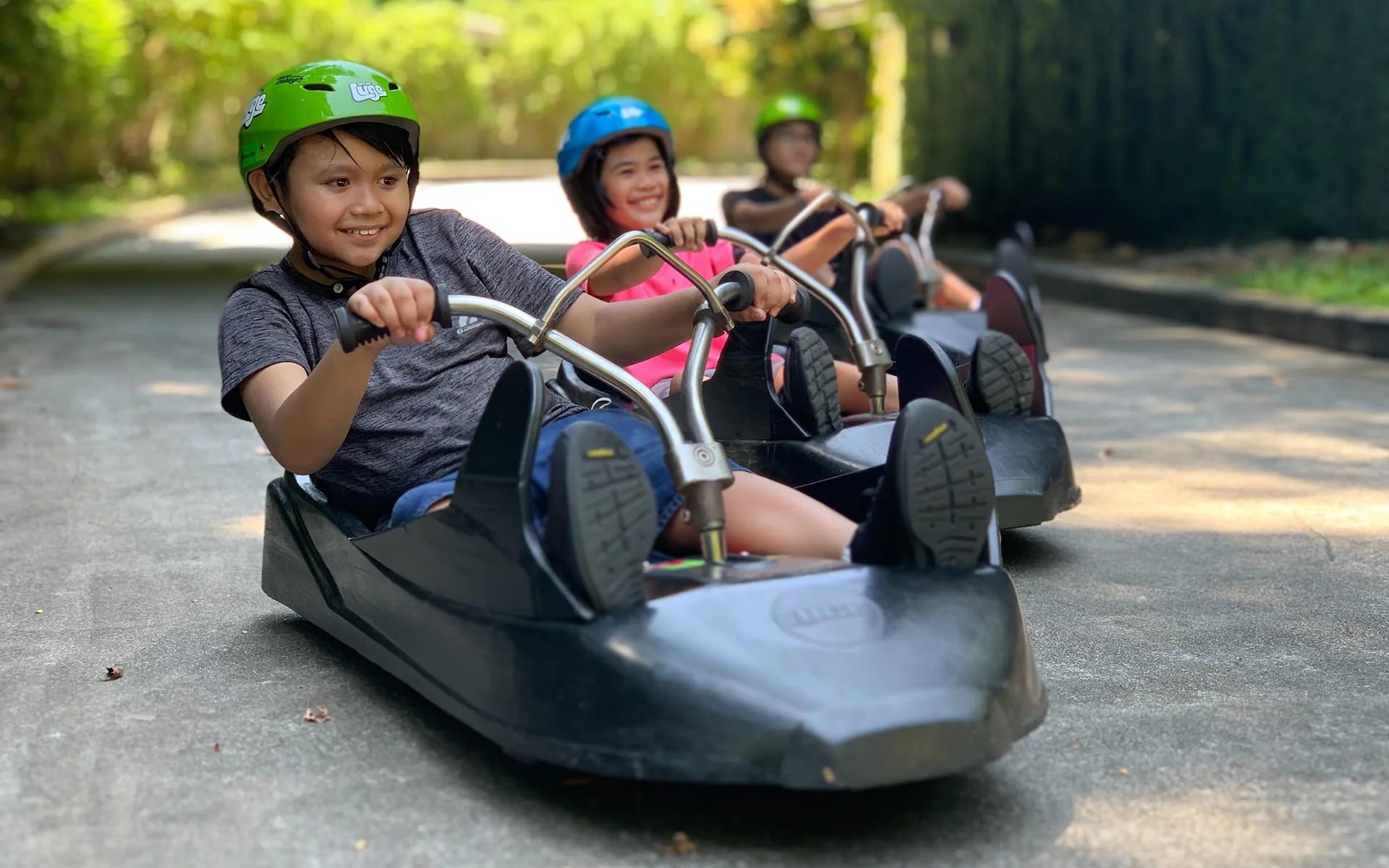 Children wearing helmets riding Skyline Luge carts along a paved track surrounded by greenery in Singapore