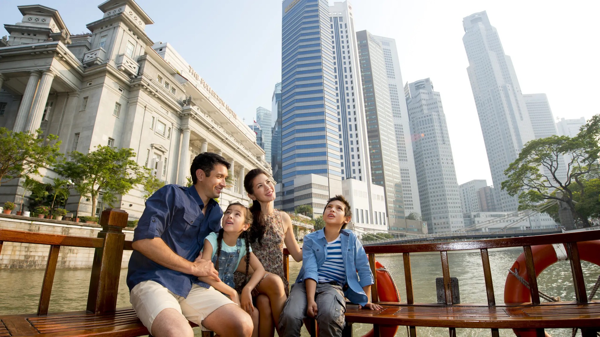 Family enjoying a Singapore River cruise with children seated on a wooden boat, looking at the city skyline and historic riverside buildings on a sunny day