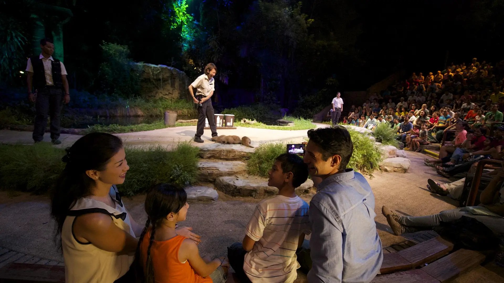 Family watching a Night Safari animal show in Singapore, with trainers on stage and a large audience seated under nighttime forest lighting.