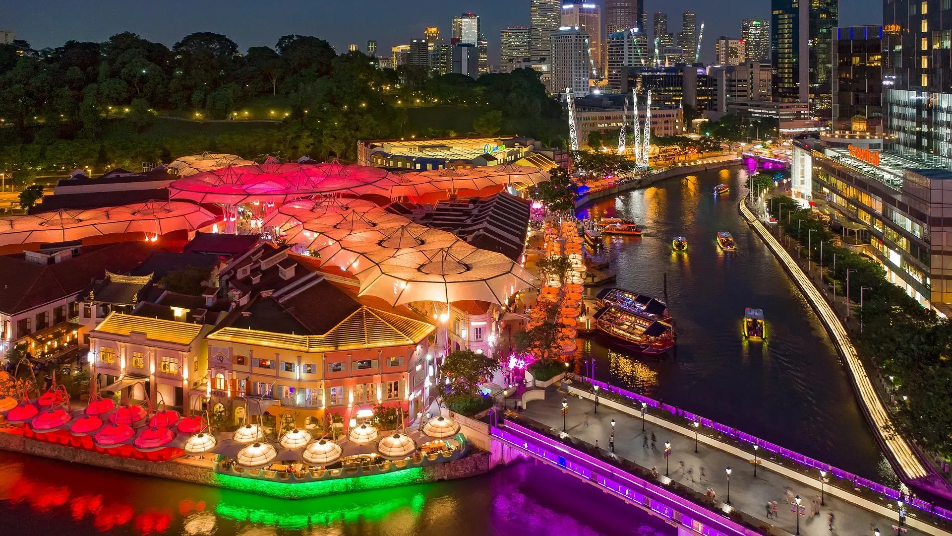 Clarke Quay riverside nightlife in Singapore with colourful illuminated buildings, neon-lit bridges, boats on the river, and the city skyline at night.