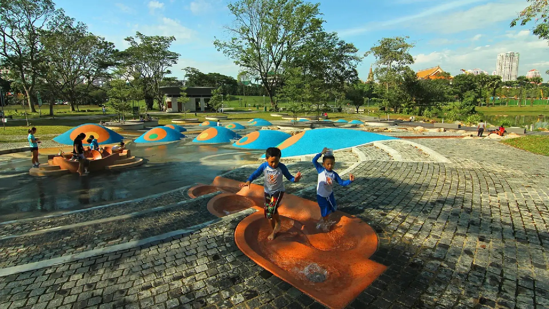 Children playing in the water playground at Bishan–Ang Mo Kio Park on a sunny day, surrounded by trees and open green space