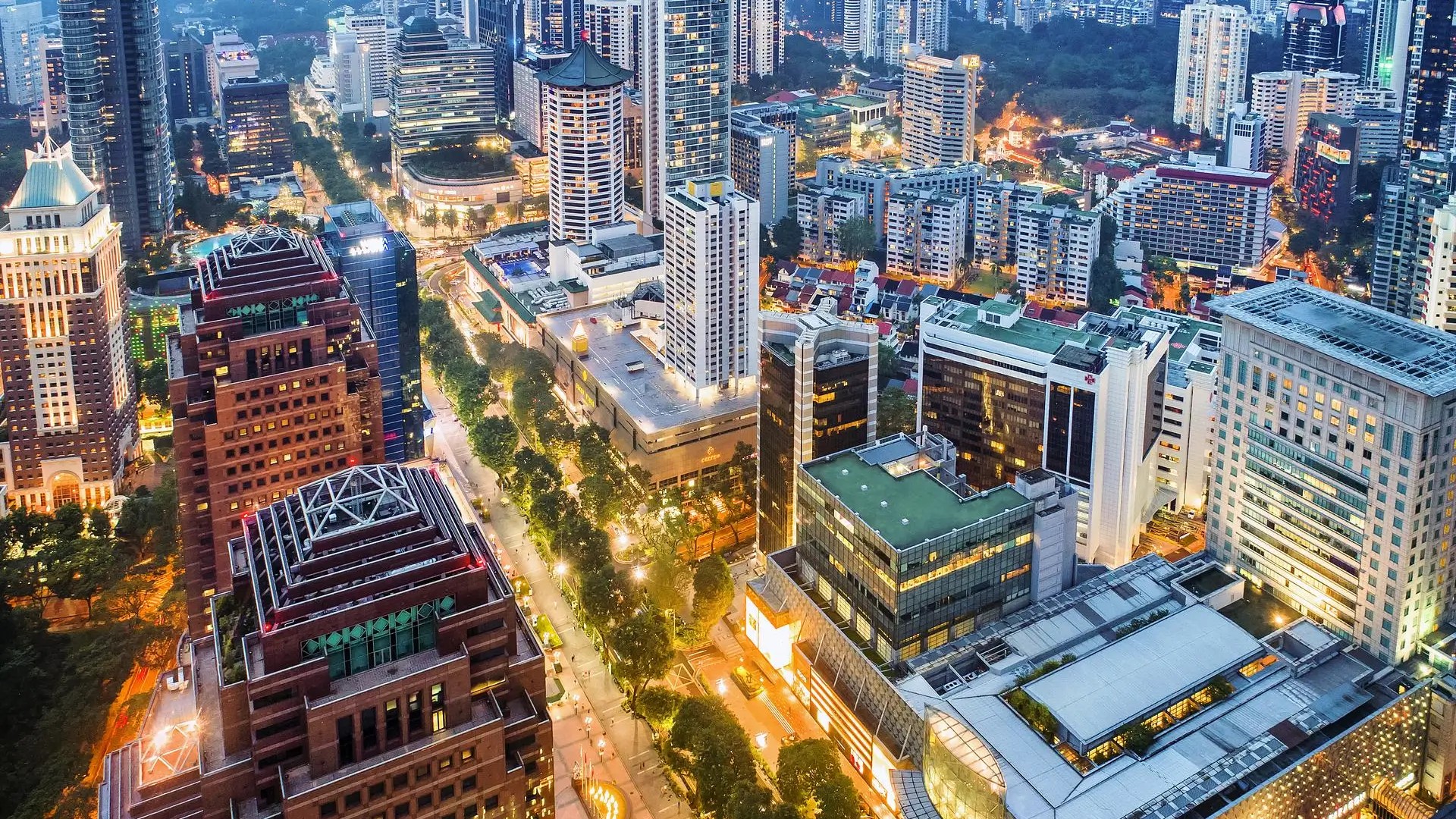 Aerial view of Orchard Road at dusk, showing illuminated skyscrapers, shopping malls, and tree-lined streets in Singapore.