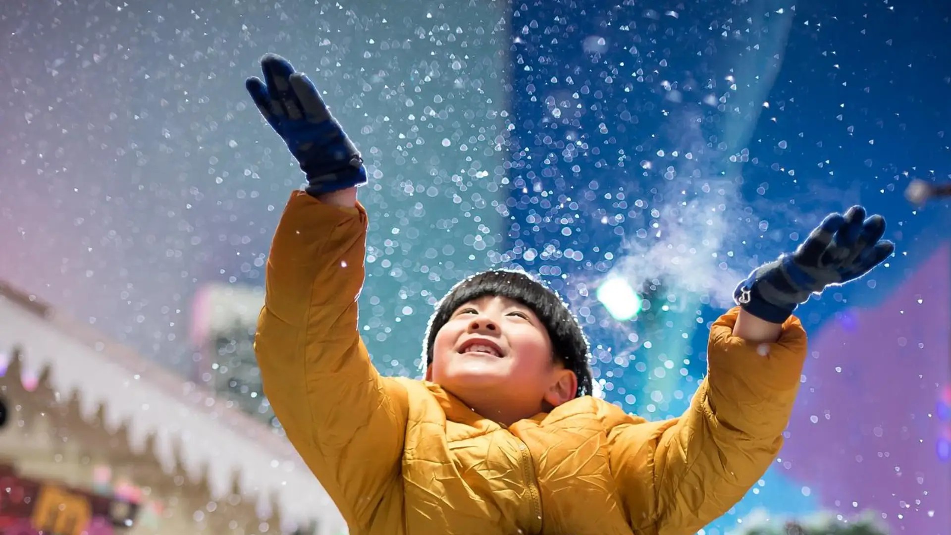Child in winter jacket reaching for falling snow inside Snow City Singapore, an indoor snow play attraction for kids.