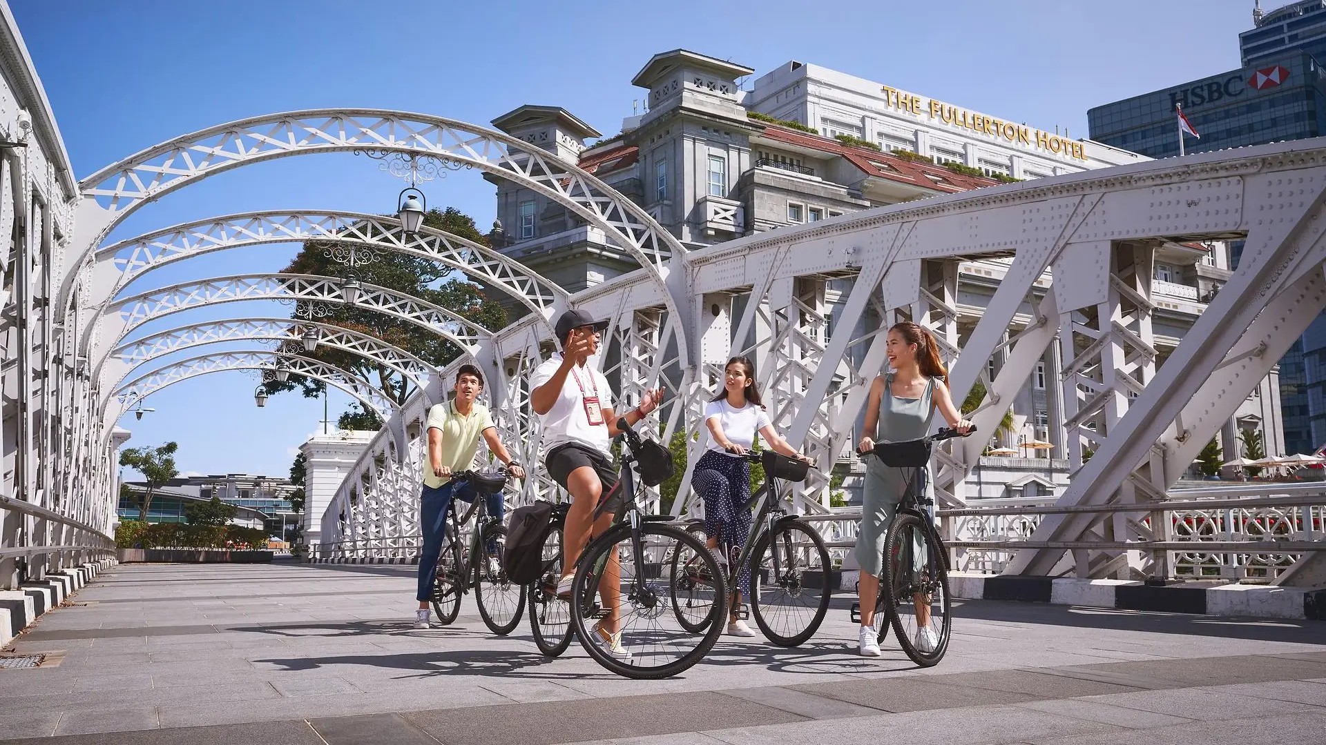 Visitors cycling across Cavenagh Bridge in Singapore’s Civic District, with The Fullerton Hotel in the background on a sunny day.