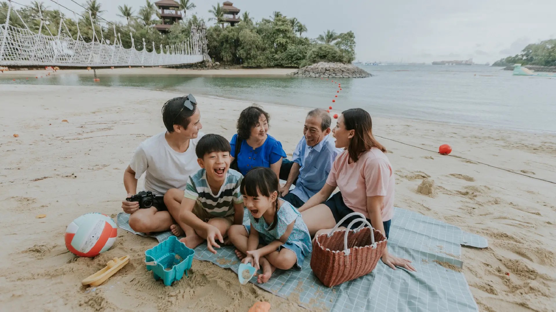 Multi-generation family enjoying a beach picnic at Sentosa Island, with children playing in the sand and grandparents relaxing by the shore.