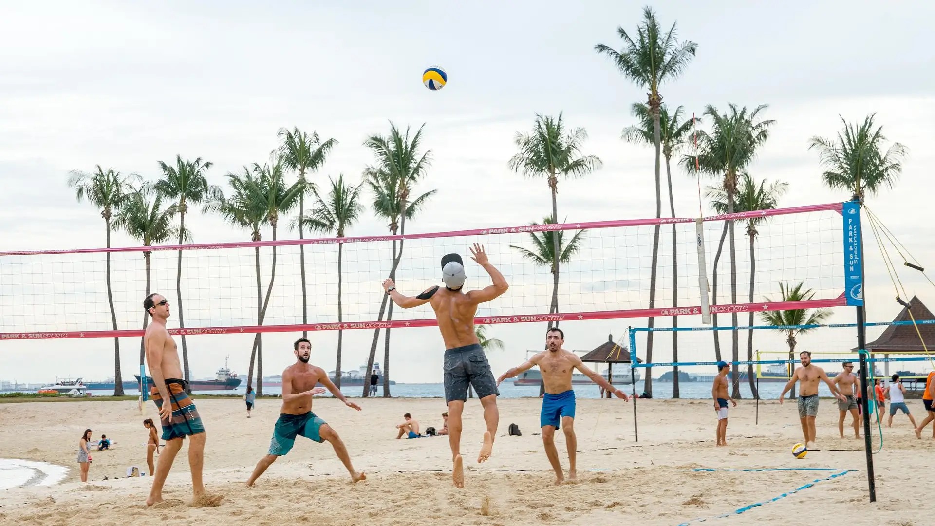 Friends playing beach volleyball on Siloso Beach, Sentosa Island, with palm trees and the sea in the background.