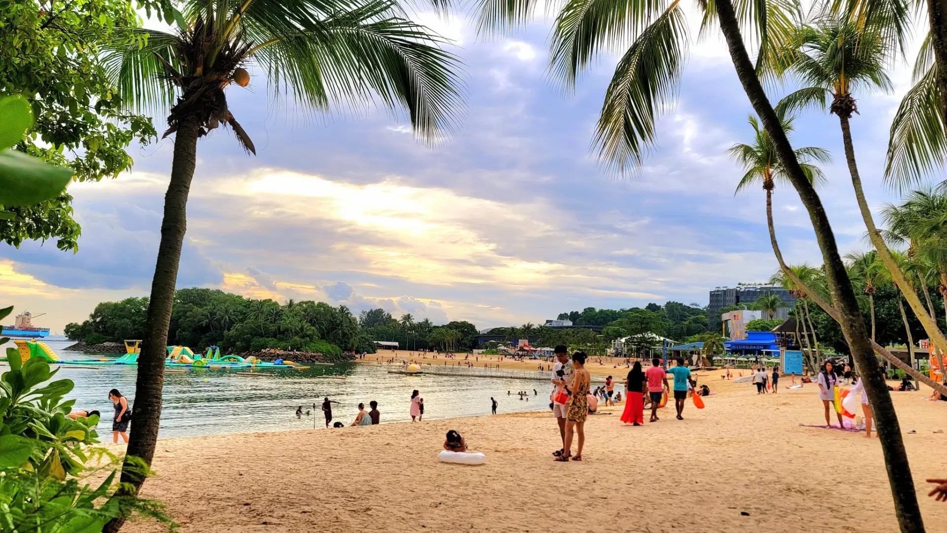 Family enjoying Palawan Beach on Sentosa Island with palm trees, calm lagoon waters, and the Southernmost Point suspension bridge in Singapore.