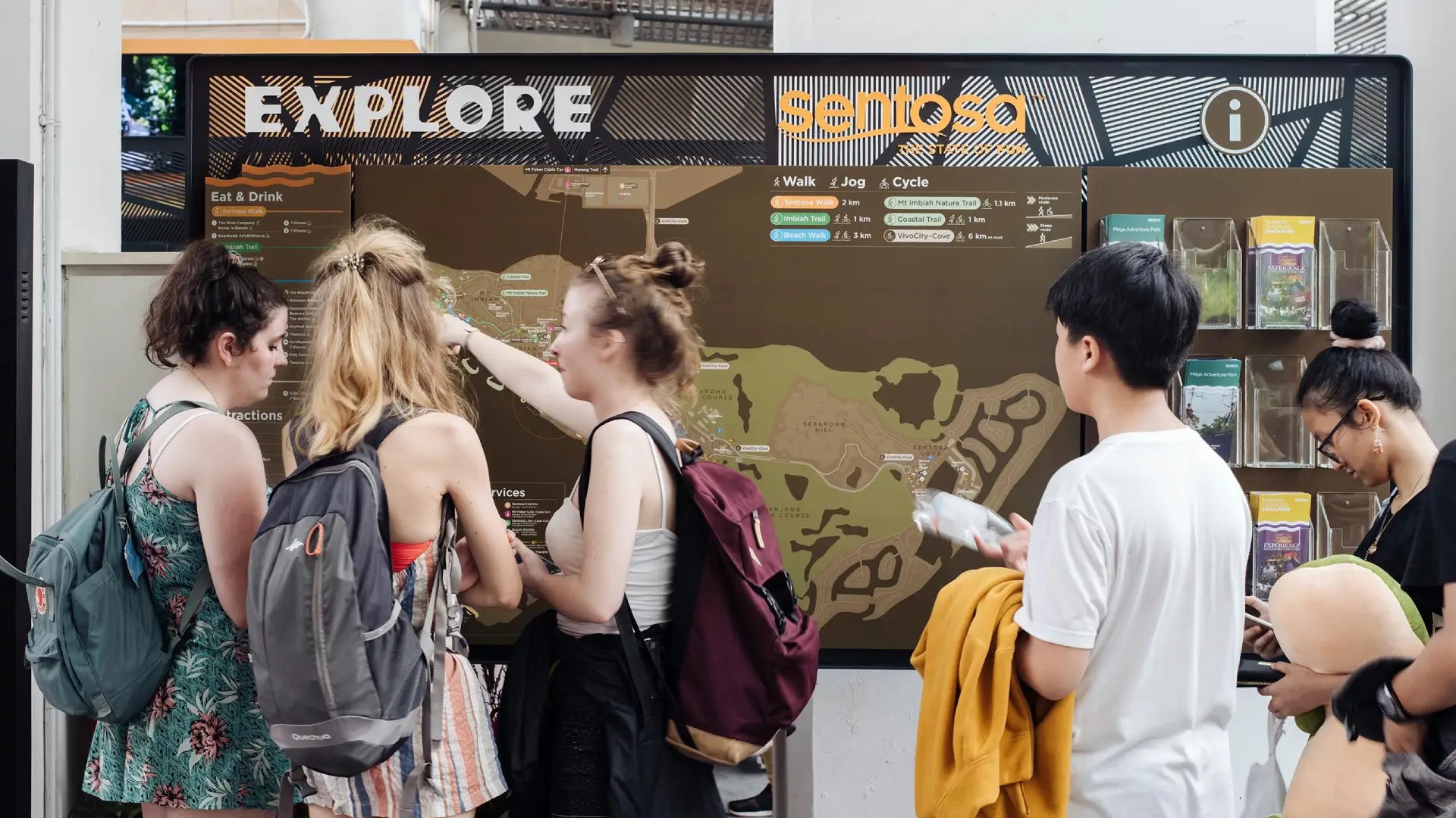 Visitors viewing a Sentosa Island map and transport information board at an information station in Singapore.