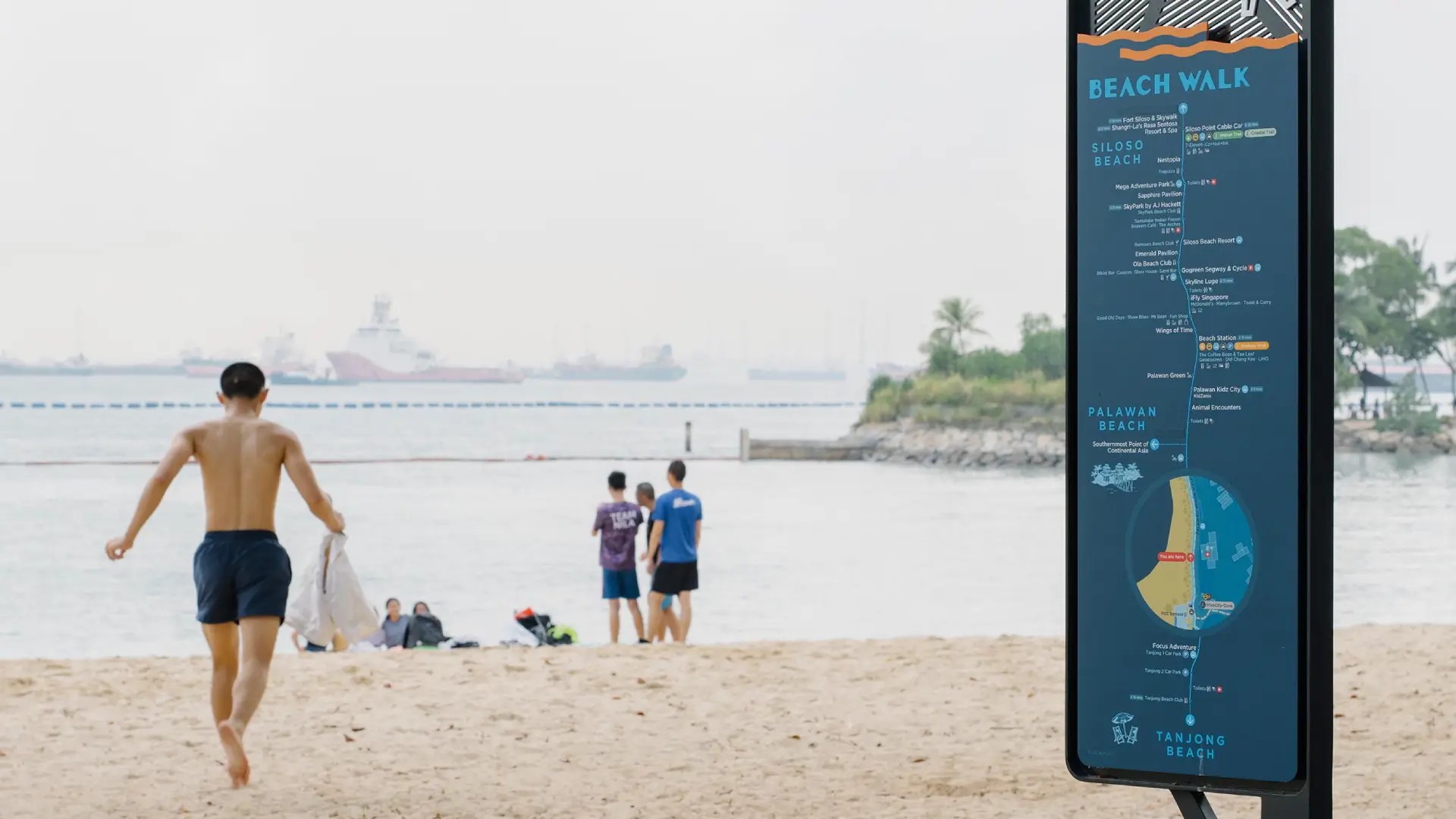 Wayfinding sign on Sentosa Beach Walk with visitors walking on the sand and coastal views in the background
