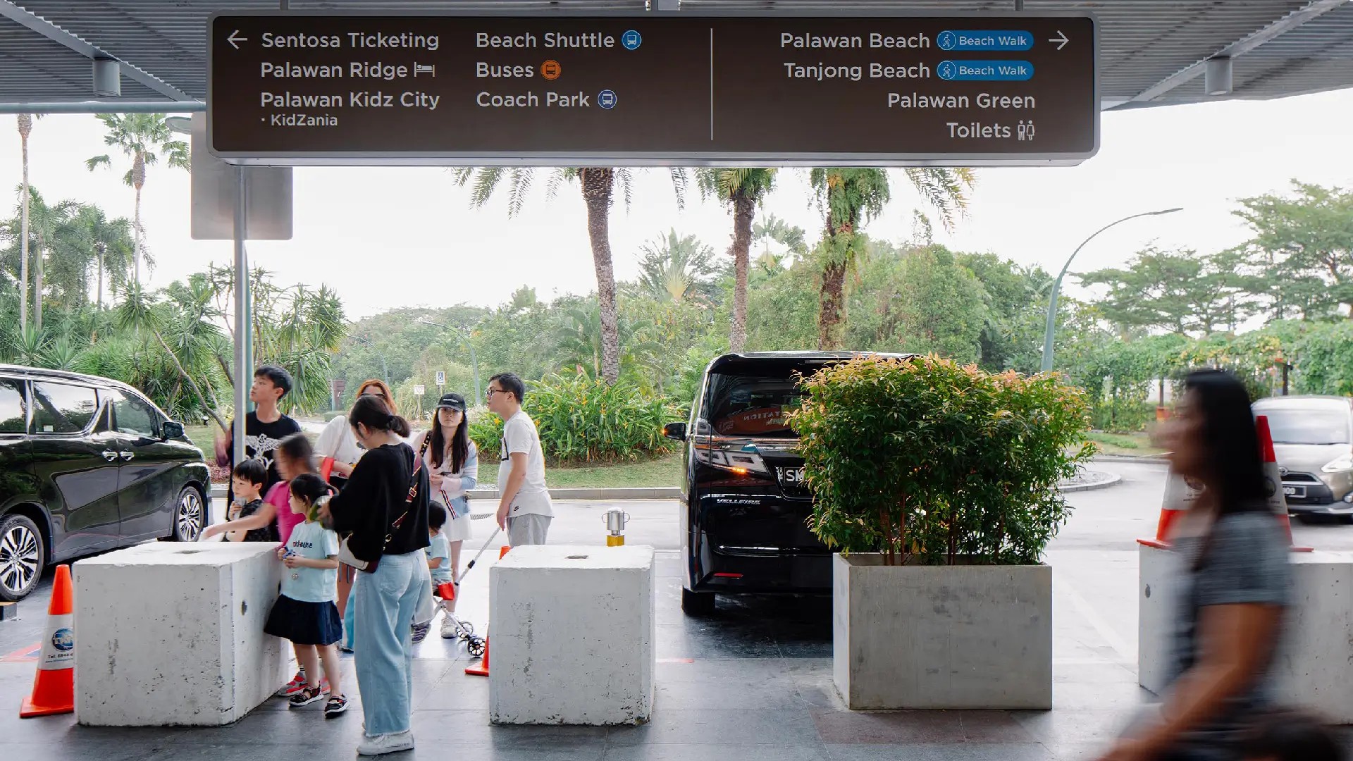 Visitors waiting at a Sentosa transport drop-off area beneath directional signage for Palawan Beach, beach shuttle buses, and key island attractions