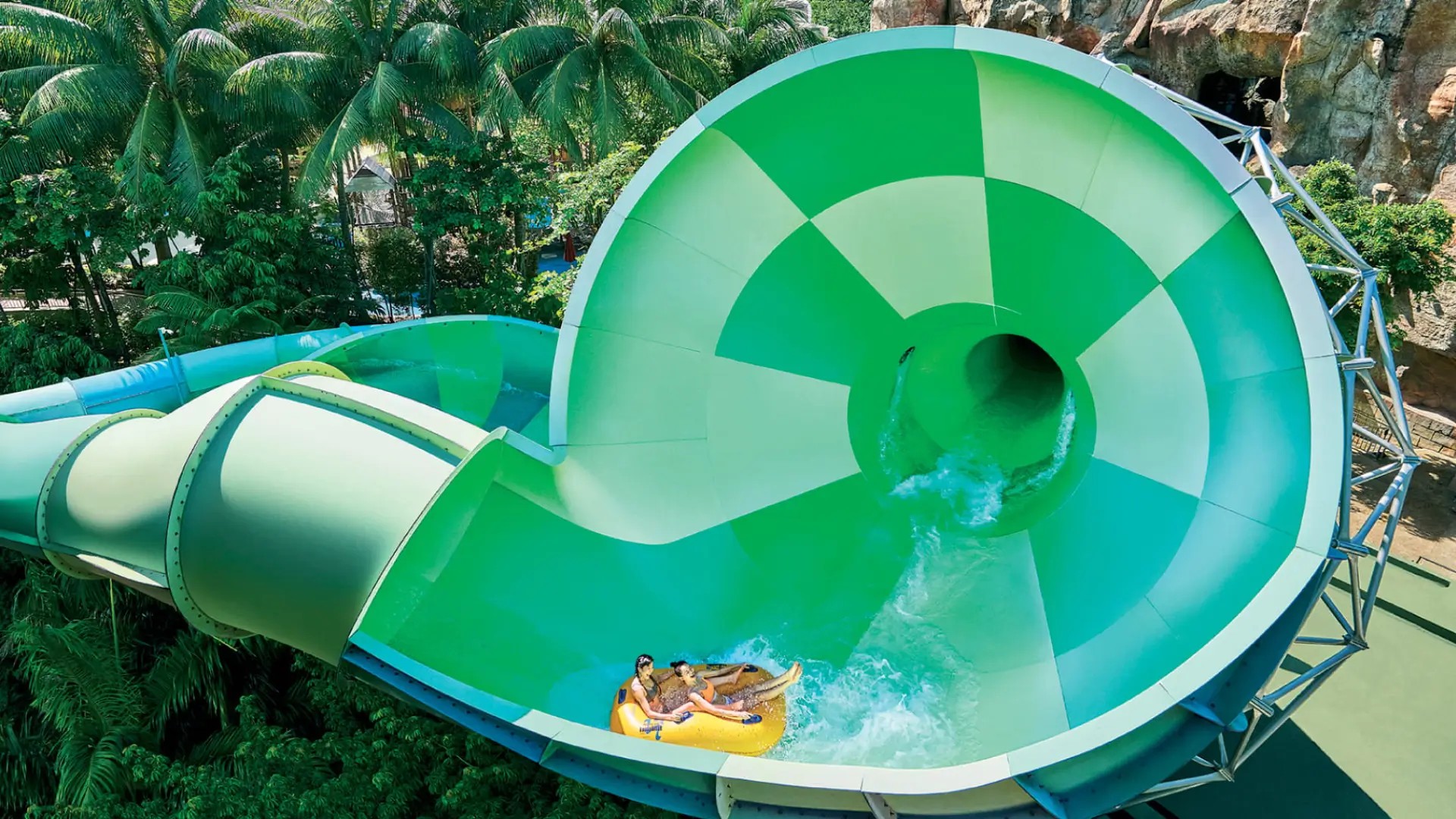 Riders on a large green spiral waterslide at Adventure Cove Waterpark on Sentosa Island, surrounded by tropical greenery