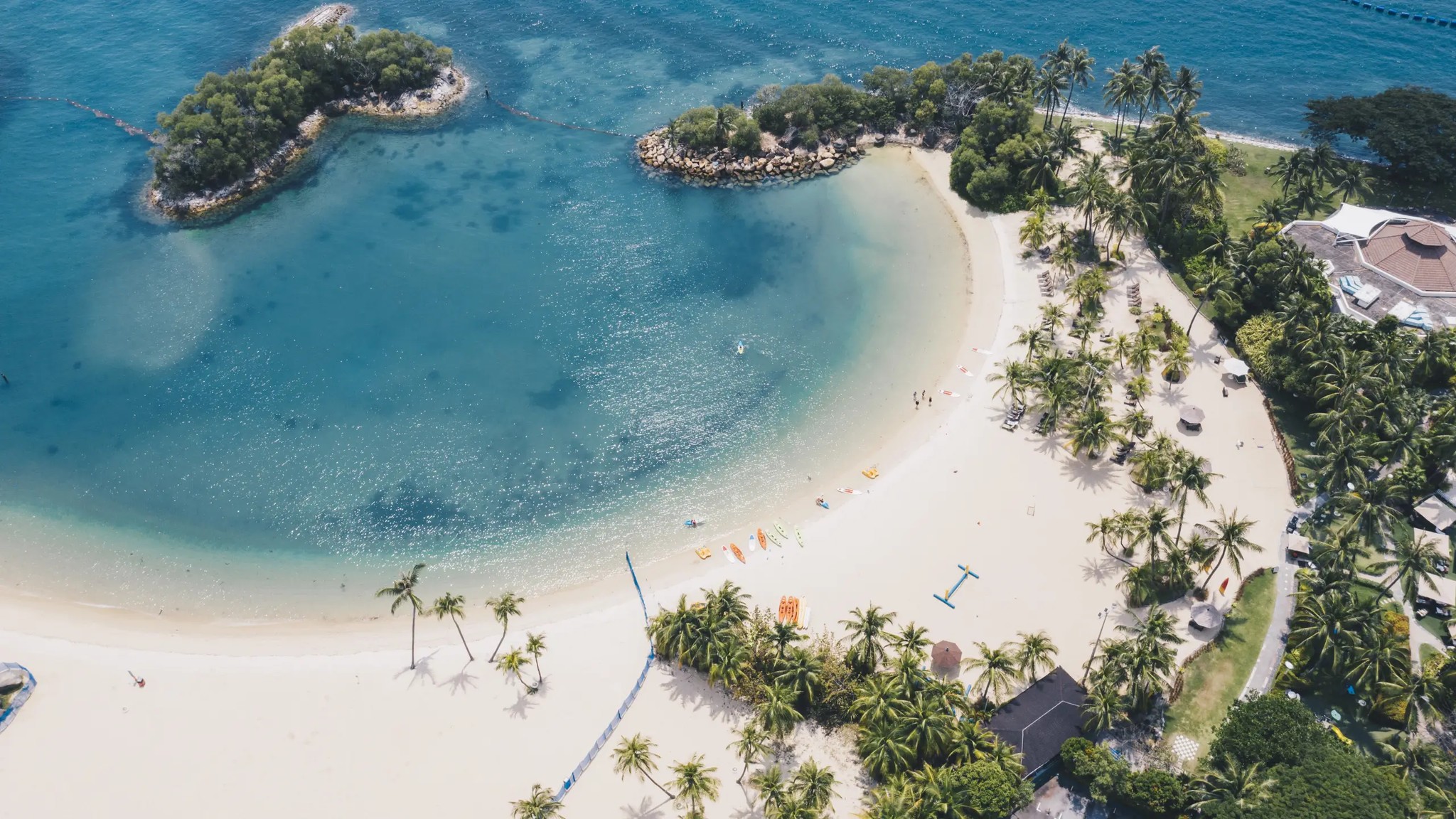 Aerial view of Siloso Beach on Sentosa Island showing a curved white-sand beach, turquoise lagoon waters, palm trees, and small offshore islets in Singapore.