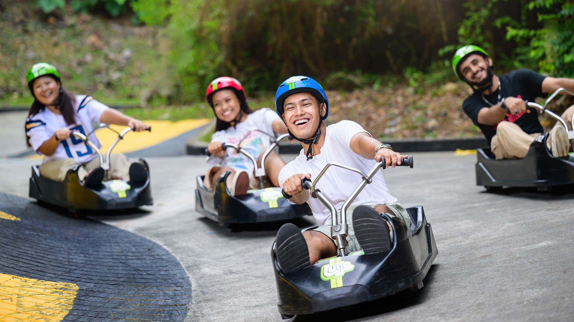 Group of friends smiling and riding Skyline Luge Singapore together as they steer their luge carts around a gentle curve on a forest-lined track.