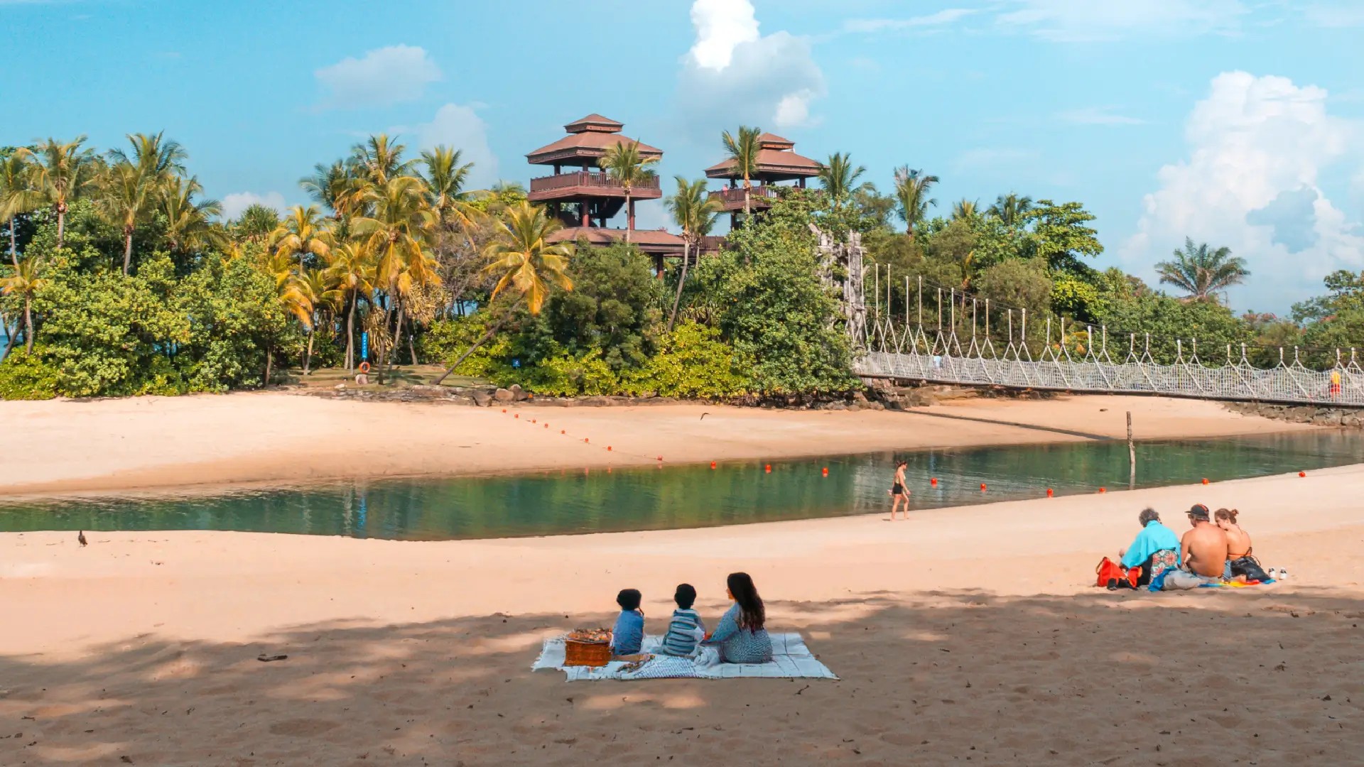 Families relaxing on Palawan Beach, Sentosa Island, with lagoon waters, palm trees and suspension bridge views