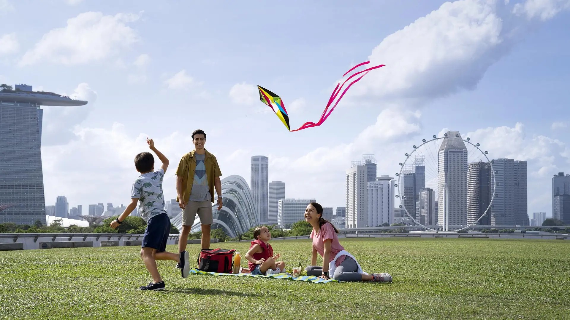 Family enjoying a picnic and flying a kite outdoors with Singapore skyline views, representing relaxed Sentosa-style attractions
