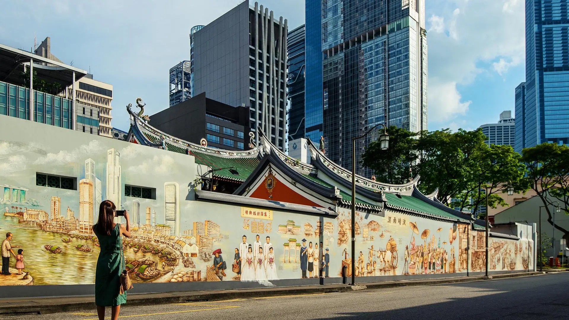 Visitor photographing a heritage mural in Singapore’s Chinatown with traditional temple architecture and modern city skyscrapers behind