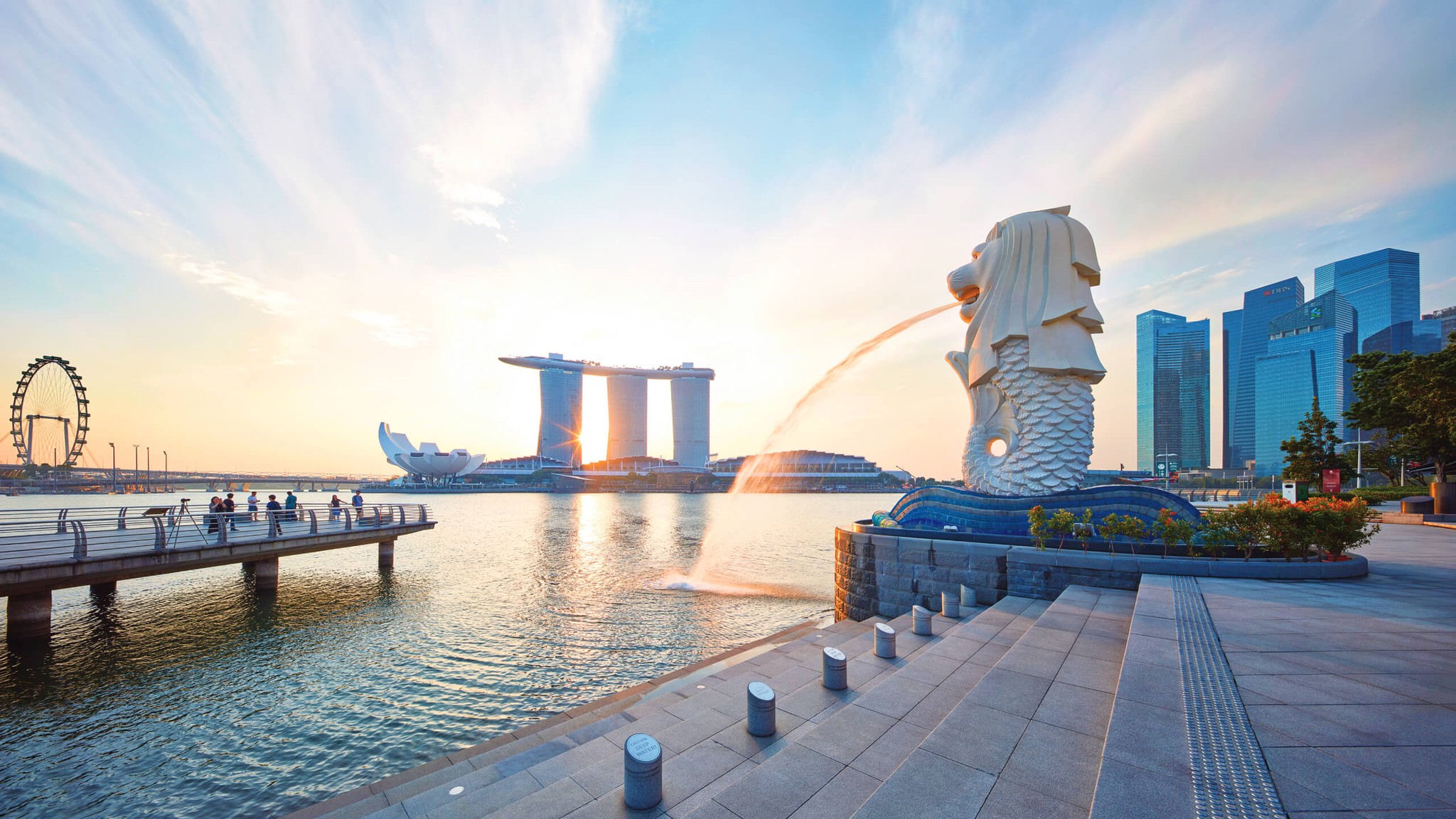 Merlion Park in Singapore at sunrise, with the Merlion statue spouting water and Marina Bay Sands and the city skyline in the background.