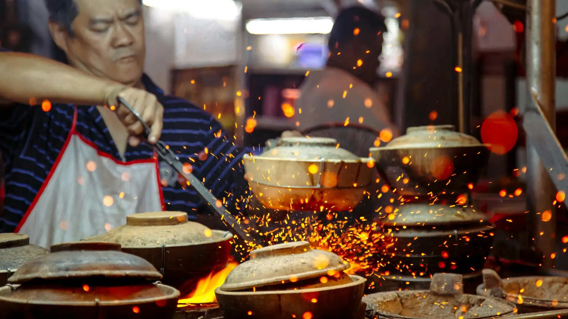 Traditional claypot cooking over an open flame with glowing sparks in a Malaysian street food kitchen.