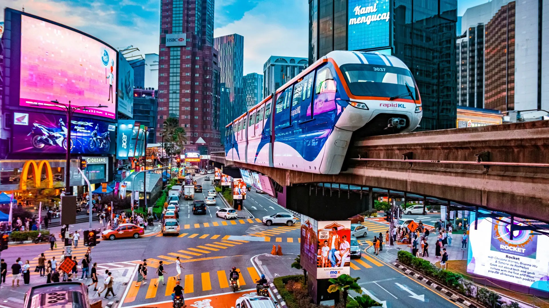 Rapid KL train travelling through central Kuala Lumpur with city skyscrapers, traffic and pedestrian crossings below.