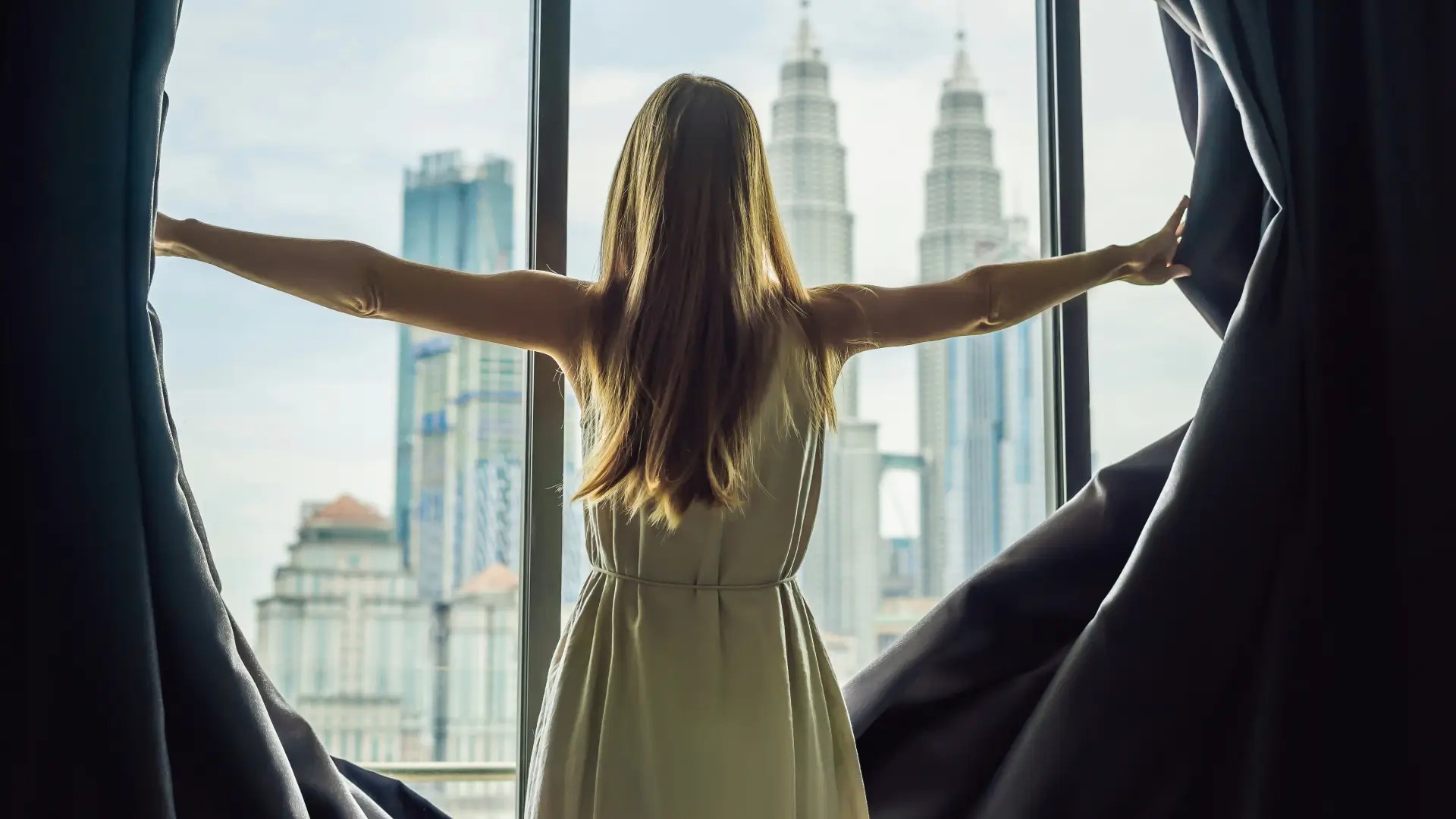 Guest standing at a hotel room window overlooking the Kuala Lumpur skyline, with the Petronas Twin Towers visible in the background.