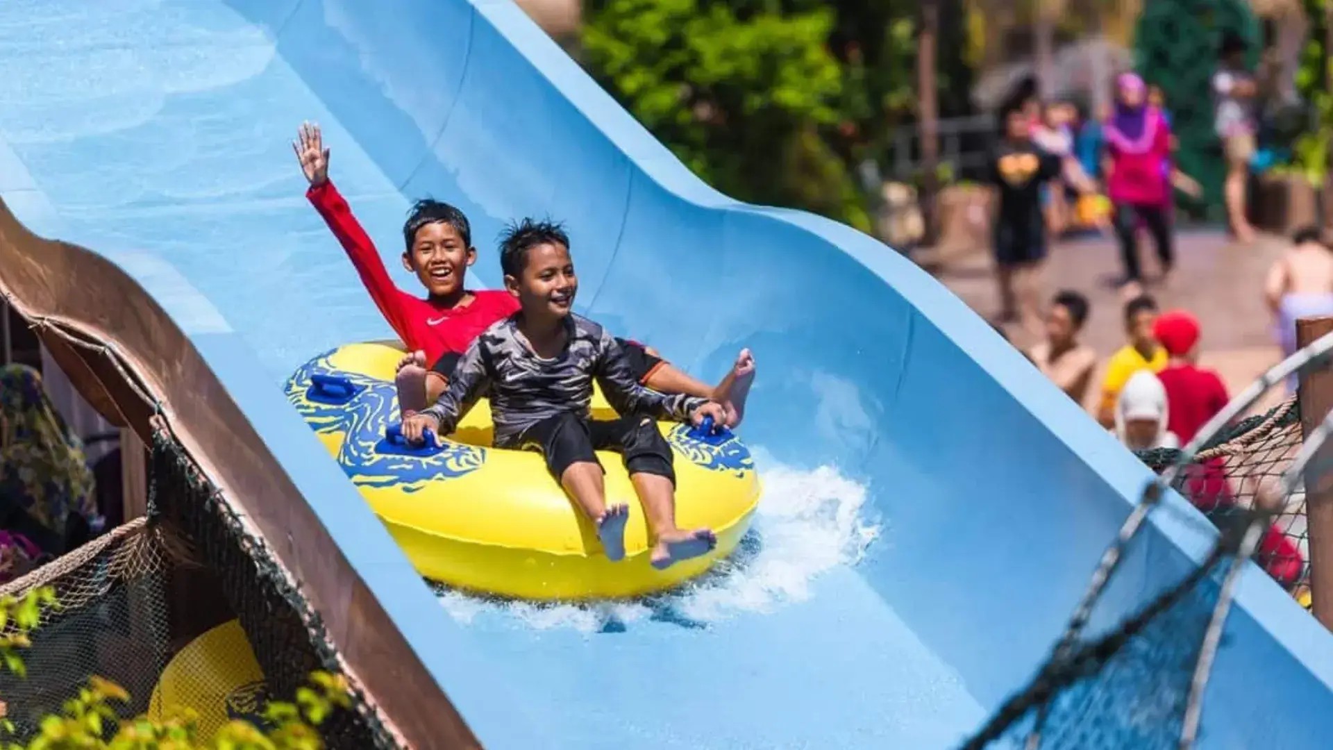Two children riding a yellow inflatable raft down a blue waterslide at SplashMania WaterPark.